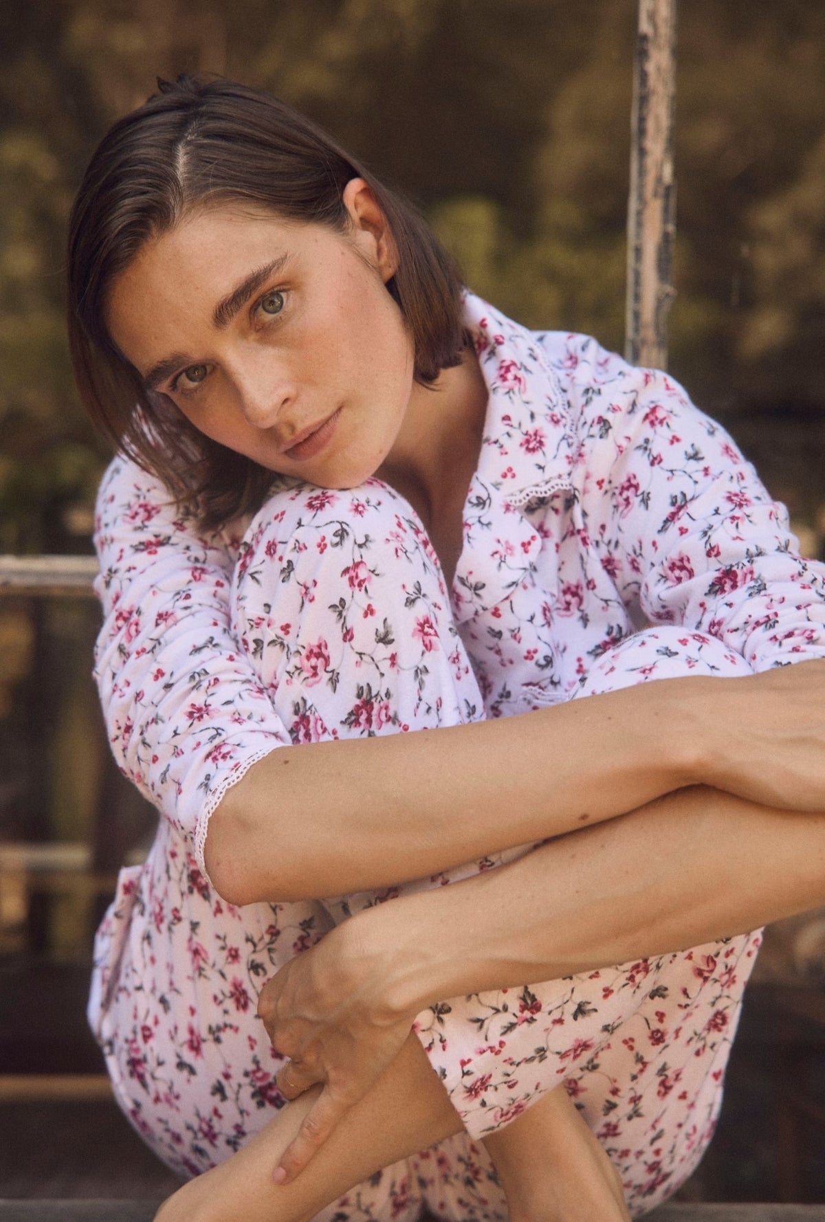 A woman with short brown hair sits cross-legged outdoors wearing a white pajama set with a pink and green floral print.