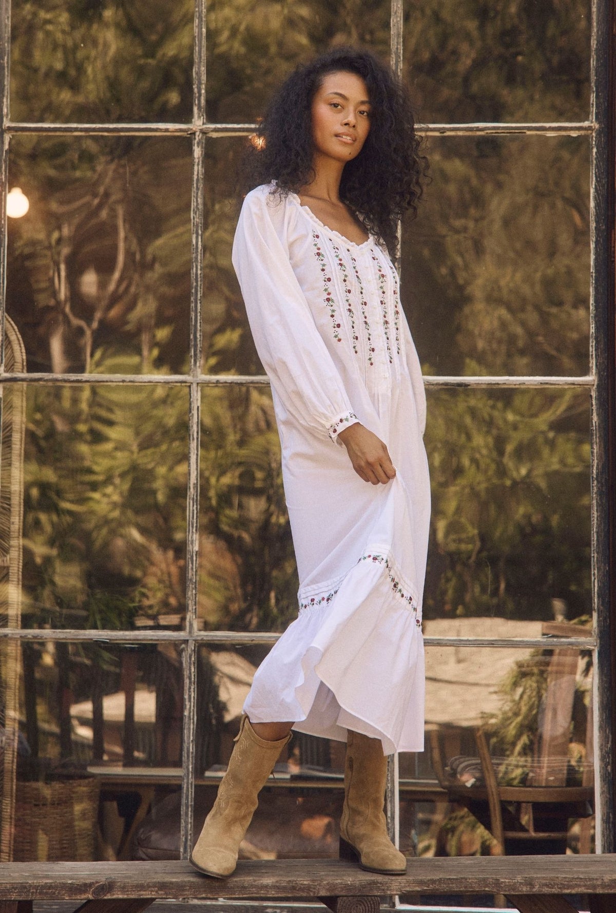 A woman wearing a white long-sleeve bohemian maxi dress with black embroidered details and tan boots, standing in front of a rustic multi-pane window with a forest background.