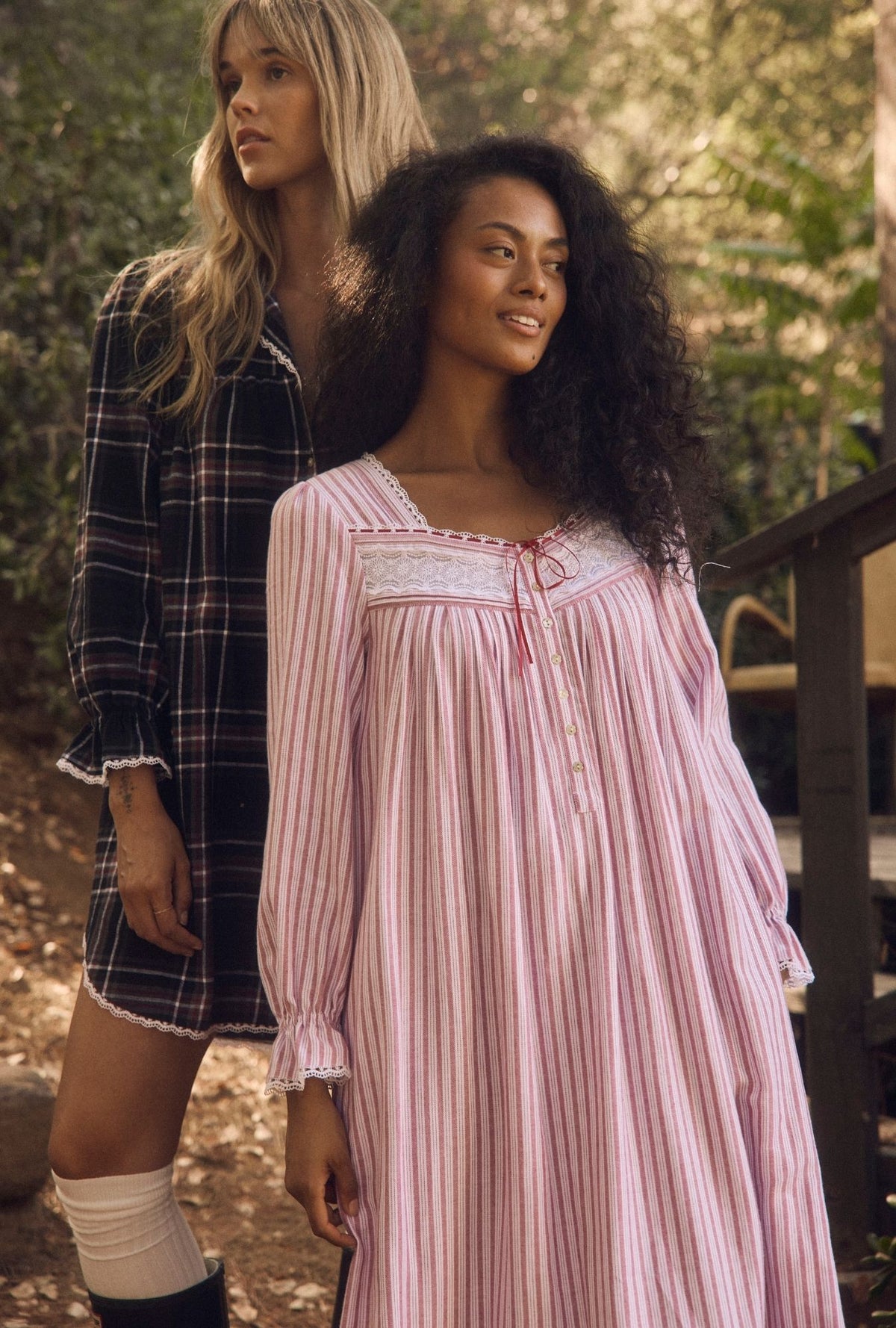 Two women standing outdoors in a wooded setting. The woman in the foreground wears a pink and white vertically striped long-sleeve nightgown with lace trim, while the woman behind her wears a dark plaid shirt dress.