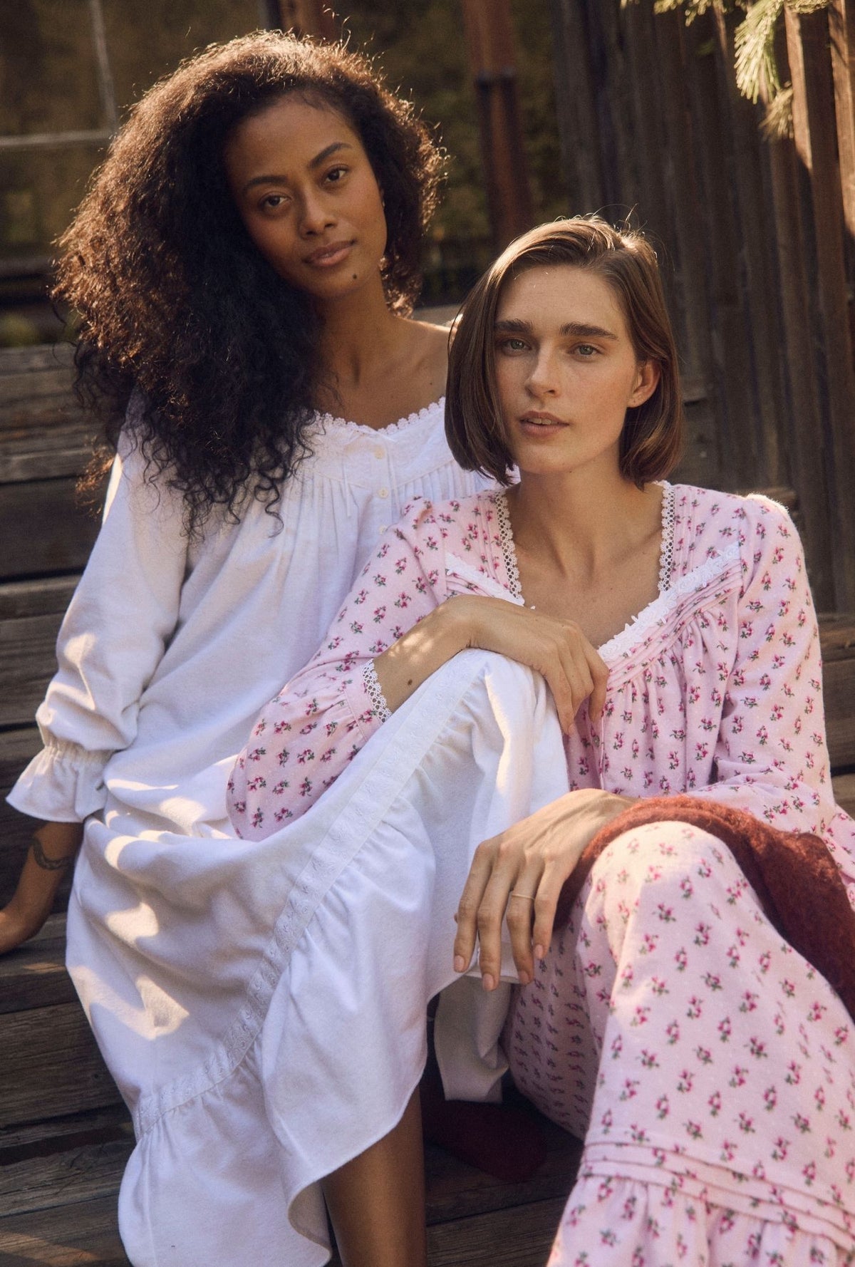 Two women sitting together outdoors in matching long-sleeve sleepwear. The woman on the left wears a loose white nightgown, and the woman on the right wears a light pink patterned pajama set with a white ruffled neckline.