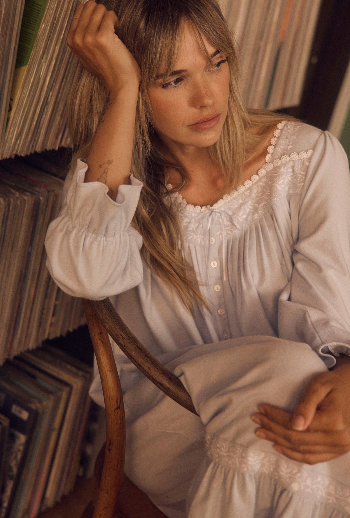 A woman with blonde hair sits in a wooden chair in front of a record rack, wearing a white long-sleeve nightgown with lace trim and white pants.