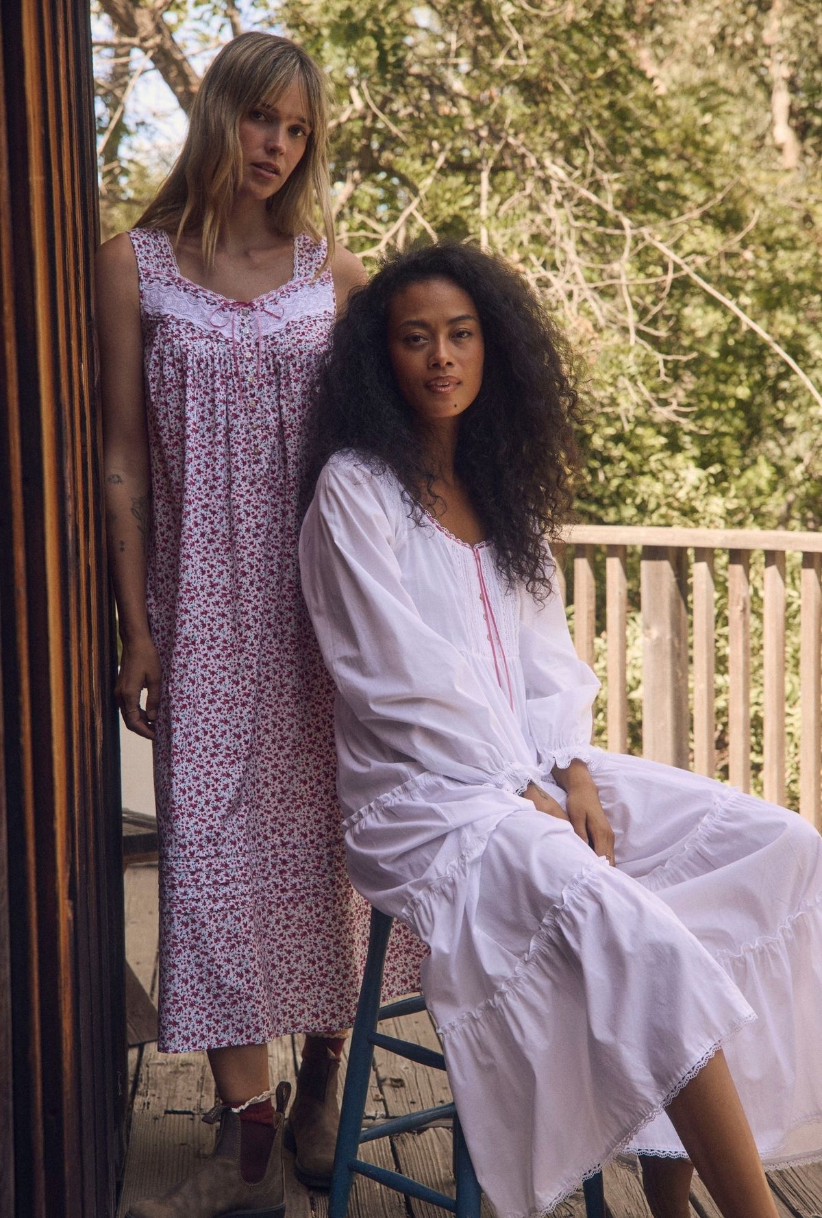 Two women modeling long sleep dresses in an outdoor porch setting. The standing woman wears a light pink and white floral print nightgown, while the seated woman wears a white long-sleeve sleep shirt dress with pink trim.