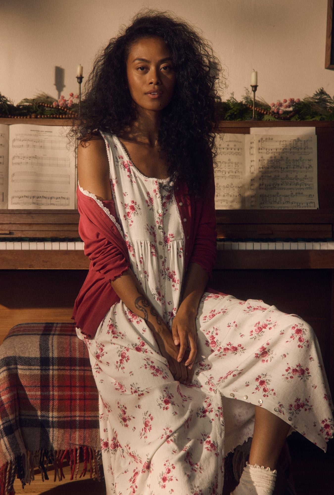 A woman with curly dark hair sits in front of a piano wearing a white floral dress with a red cardigan. Sheet music is visible on the piano, and a plaid blanket is draped over the bench.
