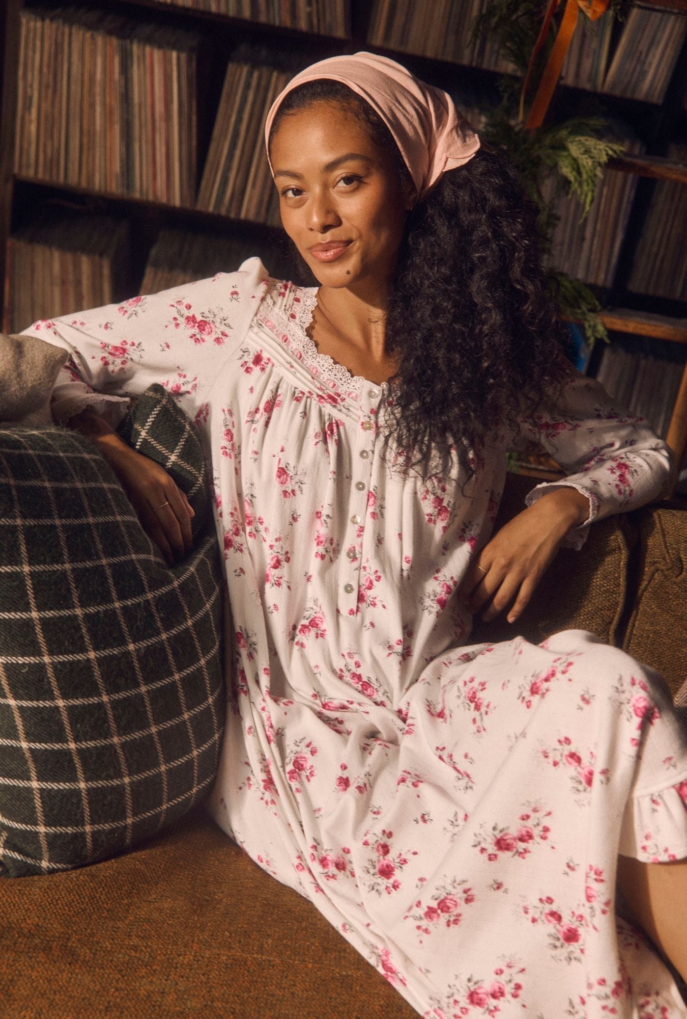 A woman wearing a long-sleeve white nightgown with a pink floral print and a matching pink headscarf, sitting in a cozy room with vinyl records in the background.